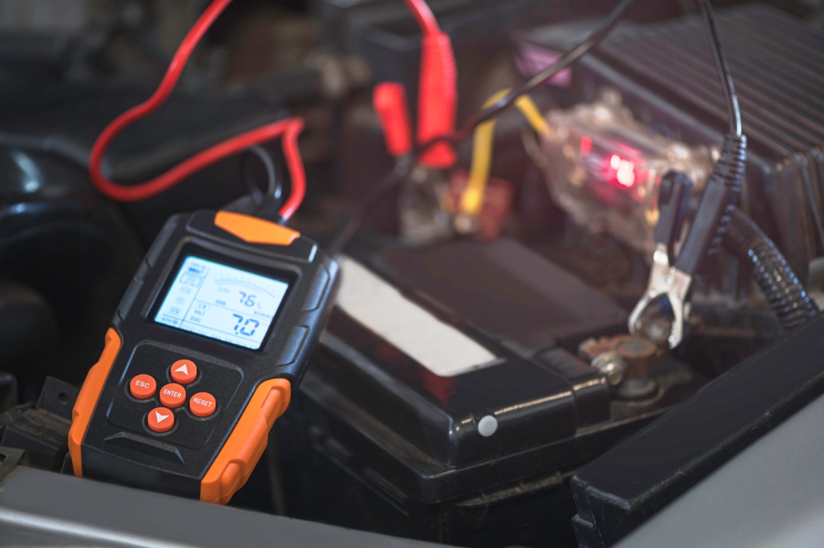 Handheld battery tester with a digital display clipped to a car battery during a charging system test.