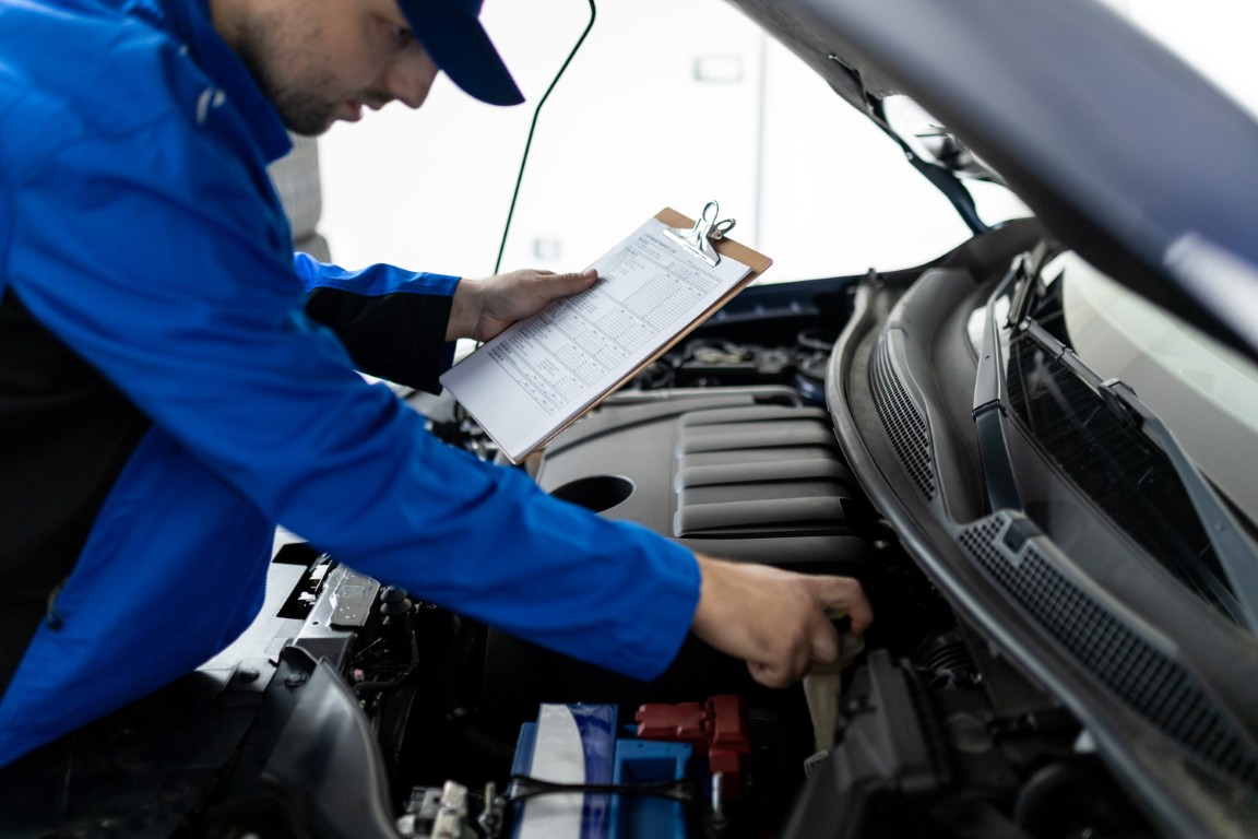 Technician filling out a multi-point inspection checklist on a clipboard beside an open hood.